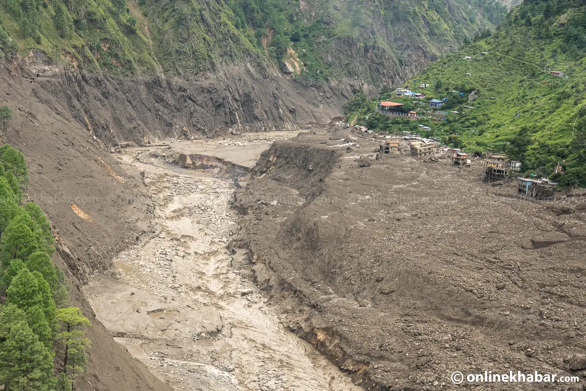 Aerial view of a debris fan of grey silt and boulders stopping at the edge of a cluster of houses