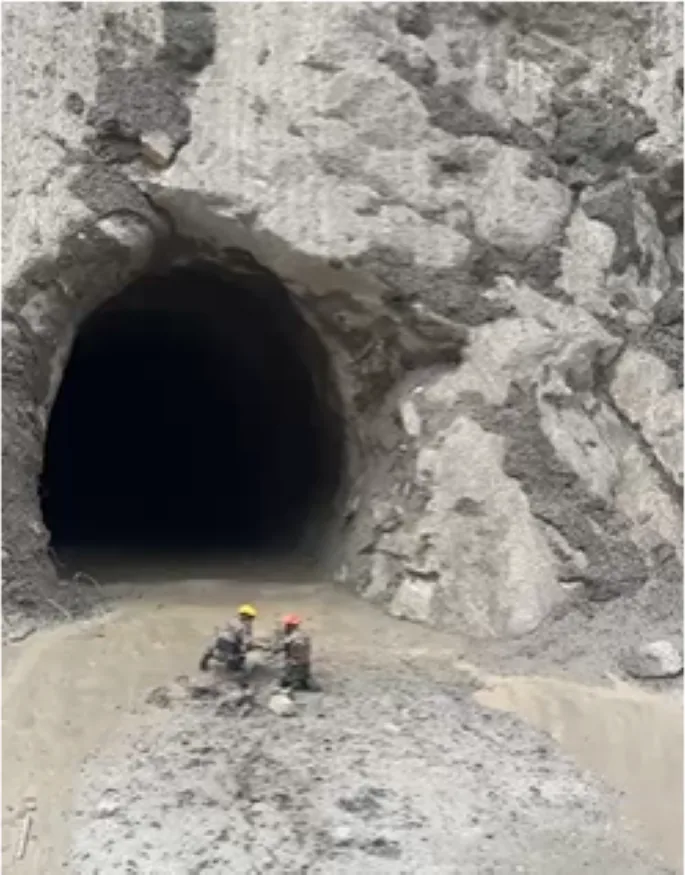 Two rescuers in hard hats kneeling at the mouth of a hydropower tunnel surrounded by grey flood silt