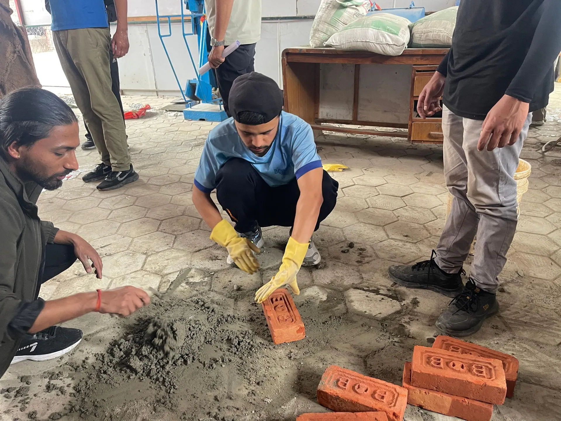 A student in gloves pressing a brick into a bed of mortar while classmates mix and watch