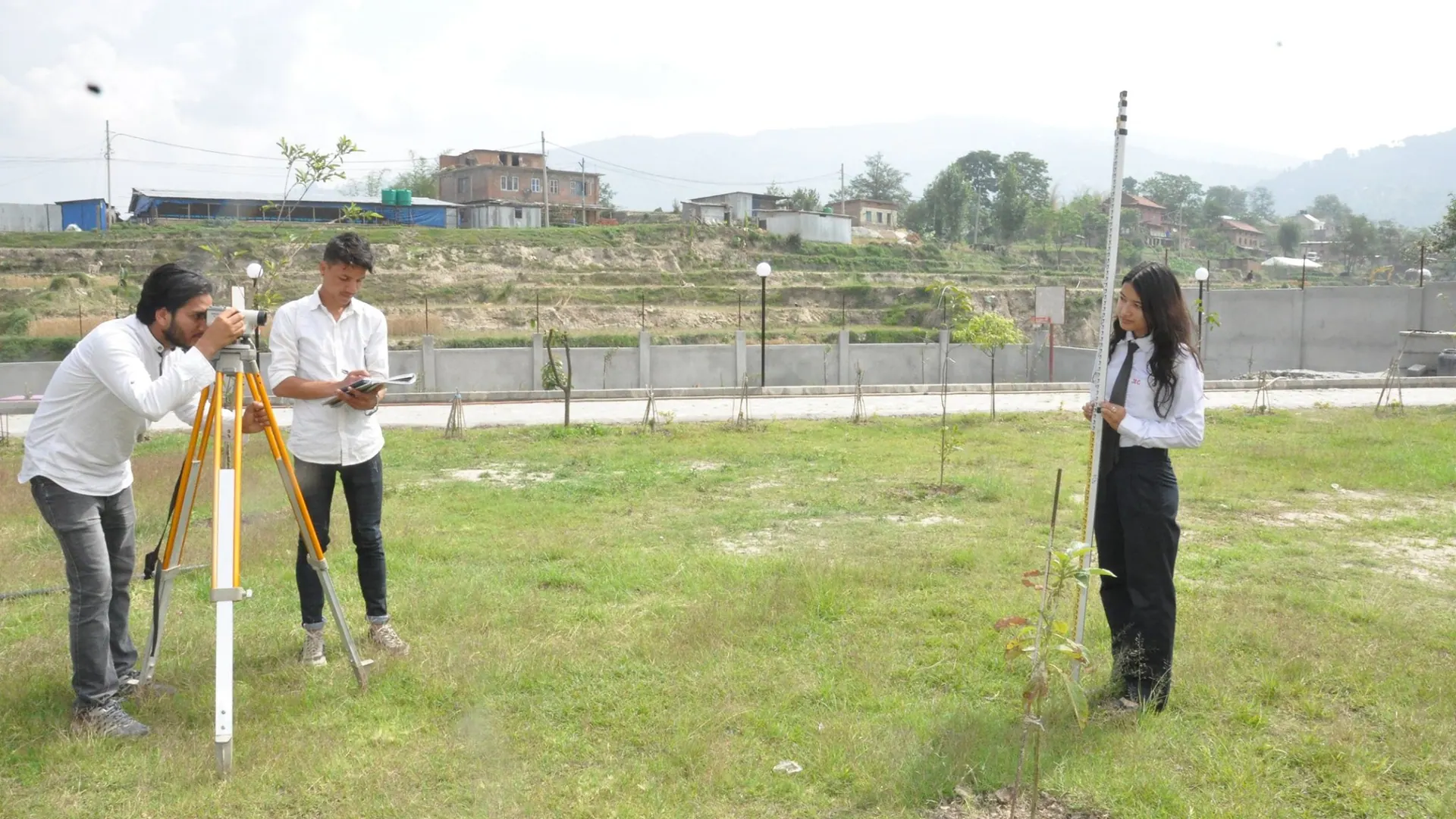 A survey party spread across open ground on the campus, one student at the instrument and another holding the staff
